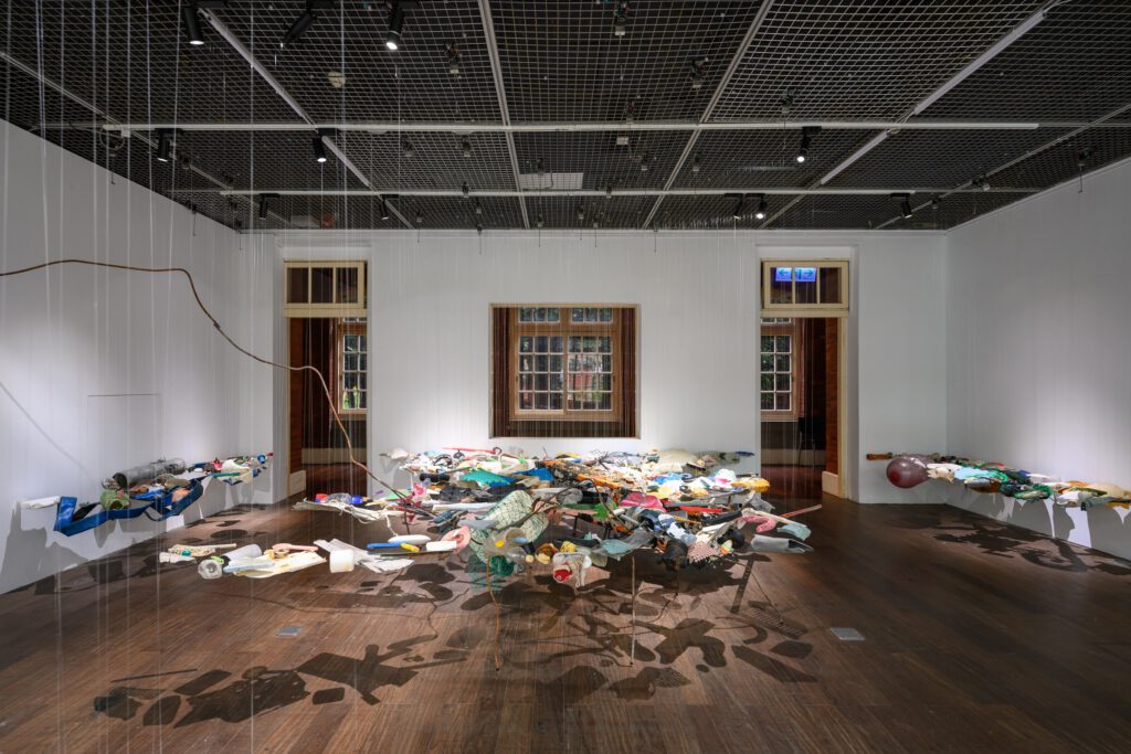 Suspended assemblage of colourful objects casting shadows across a gallery floor.
