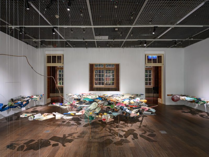 Suspended assemblage of colourful objects casting shadows across a gallery floor.