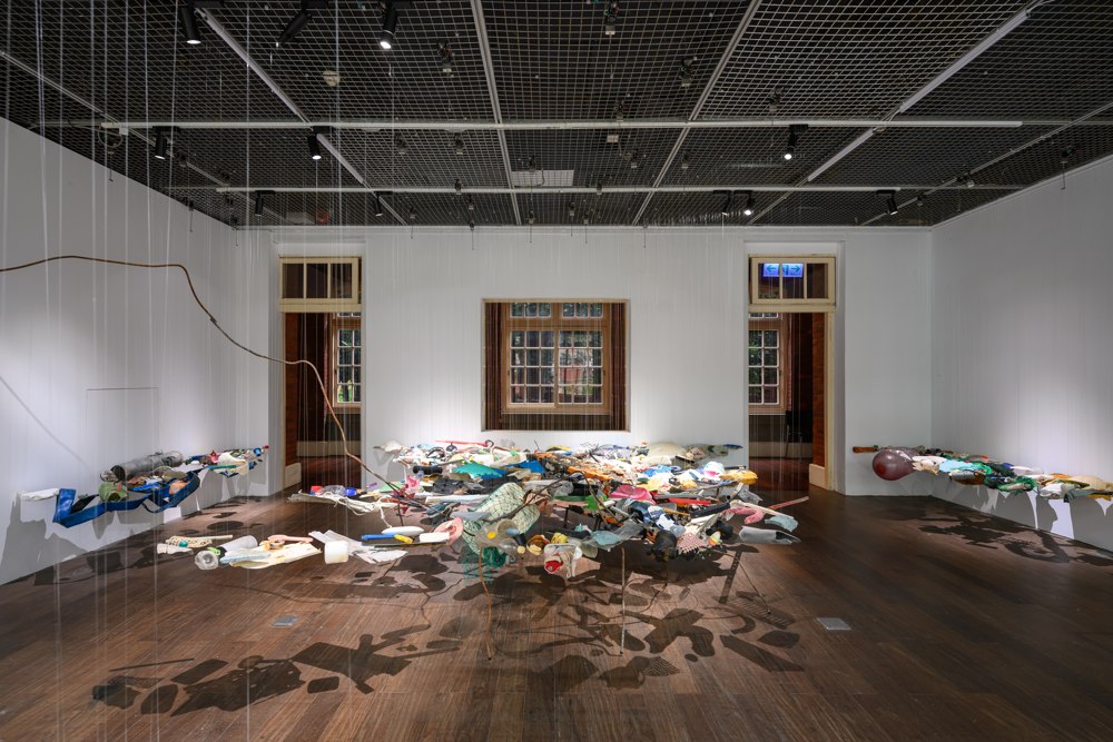 Suspended assemblage of colourful objects casting shadows across a gallery floor.